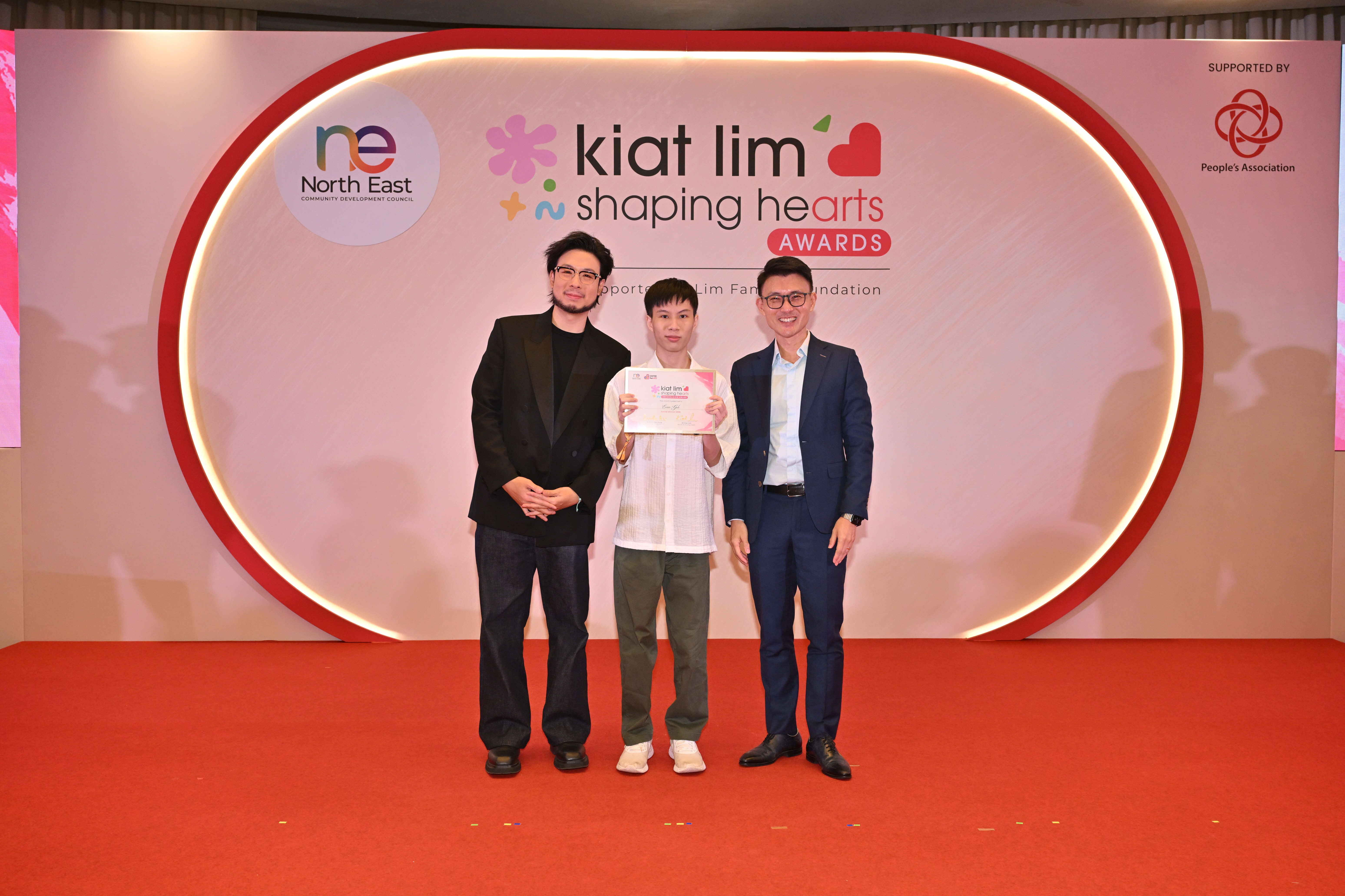Award recipient standing on stage with the presenter, holding their certificate and posing for a group photo during the award ceremony, with a backdrop reading ‘Kiat Lim-Shaping Hearts Awards'.